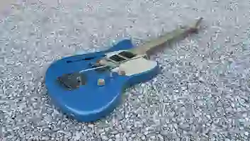 Eine handgefertigte blaue semiakustische E-Gitarre mit cremefarbenem Schlagbrett liegt auf hellem Kies